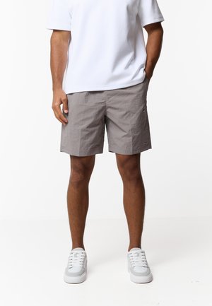 Mężczyzna w jasnoszortach casual, białych trampkach i białej koszulce z krótkim rękawem stoi z jedną ręką w kieszeni.