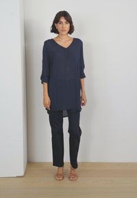 Tunic blu navy con scollatura a V e maniche a tre quarti, abbinata a pantaloni scuri e sandali, su uno sfondo neutro.