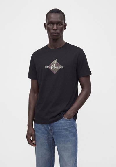 Άνδρας που φοράει μαύρο t-shirt Emporio Armani με λογότυπο, μπλε τζιν, στέκεται με το ένα χέρι στην τσέπη μπροστά σε απλό ανοιχτό φόντο.