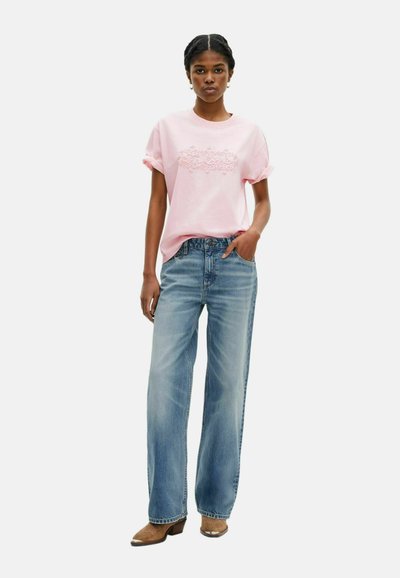 Camiseta rosa claro con gráfico en relieve, mangas arremangadas; combinada con pantalones vaqueros de pierna ancha de mezclilla azul y botas de tacón marrones.