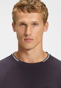 Dark purple T-shirt with a round neckline, featuring a contrasting white stripe along the collar and a subtle logo detail on the chest.