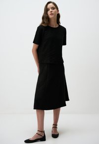 Schwarzes Zweiteiler-Outfit aus strukturiertem Stoff. Kurzärmliges Oberteil mit Rundhals-Ausschnitt, schwingender knielanger Rock, kombiniert mit schwarzen Ballerinas.