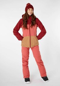 Roze en kastanjebruine geïsoleerde jas met een beige paneel, gecombineerd met bijpassende roze sneeuwbroek en een bordeauxrode gebreide beanie met pompon. Zwarte laarzen.