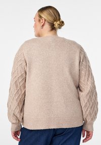 Bežs adīts džemperis ar teksturētām, kabelveida piedurknēm. Tam ir brīvs piegriezums un ribota apakša. Mīksts materiāls ar smalku, mājīgu izskatu.