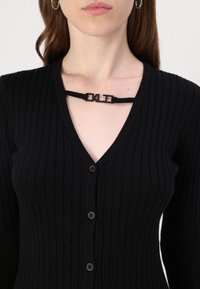 Fekete bordás kardigán, V nyakkivágással, öt gombbal, és egy fekete szalaggal, amely az "OKL" felirattal díszíti a nyakkivágást.