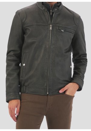 Mand iført en sort læderjakke med lynlås foran, lynlåslomme på brystet, knaplukning på sidelommerne og brune bukser mod en ensfarvet baggrund.