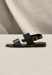 Μαύρο επίπεδο σανδάλι με δύο φαρδιές λουριά και χρυσή αγκράφα σε λουράκι αστραγάλου, τοποθετημένο σε ουδέτερη υφή επιφάνειας και φόντου.