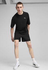 Černé bavlněné tričko a černé šortky, s malým bílým logem Puma, v kombinaci s bílými teniskami a pruhovanými ponožkami. Jednoduchý, moderní design.