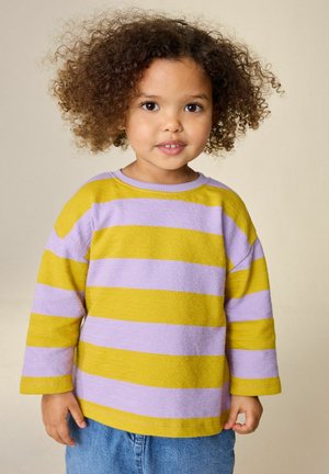 Young child with curly hair wearing a yellow and lavender striped sweater and blue jeans, standing against a neutral background.