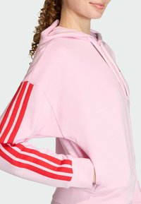 Ružová mikina s kapucňou, ktorá má červené pruhy na rukávoch, jemnú látku, voľný strih a dizajn s klokankou.