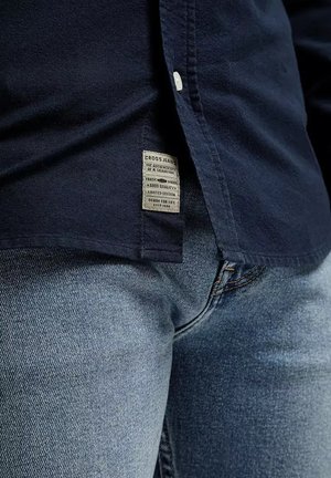 Granatowa koszula zapinana na guziki z metką materiałową na dole, w połączeniu z jasnoniebieskimi dżinsami o klasycznym kroju i delikatnej teksturze.