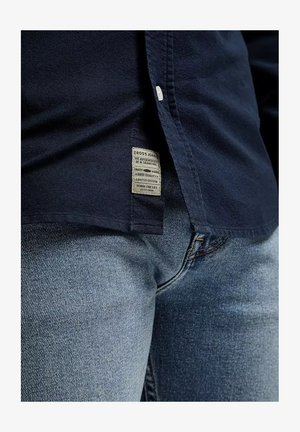 Granatowa koszula zapinana na guziki z metką materiałową na dole, w połączeniu z jasnoniebieskimi dżinsami o klasycznym kroju i delikatnej teksturze.