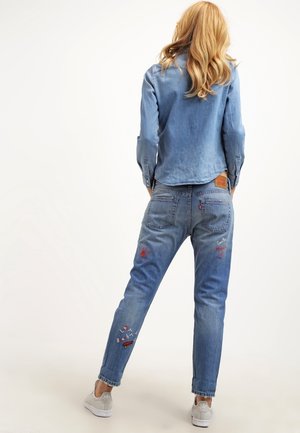 Femme aux longs cheveux blonds portant une chemise en jean bleu clair et un jean bleu brodé, debout de dos sur un fond uni.