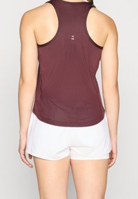 Maroonfarbenes ärmelloses Sporttanktop mit Mesh-Struktur, mit abgerundetem Saum, minimalistischem Rücken-Design, kombiniert mit weißen Shorts.