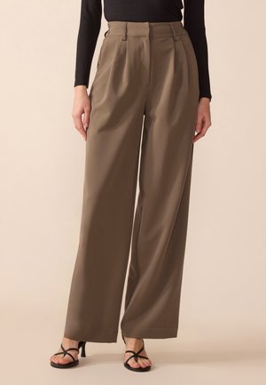 Vrouw draagt taupekleurige, hoog getailleerde, wijd uitlopende broek, zwarte top met lange mouwen en zwarte sandalen met hak en bandjes op een beige achtergrond.