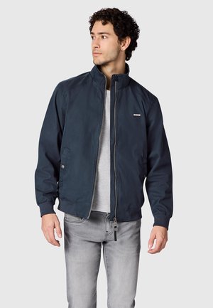 Mand iført en marineblå lynlåstrøje over en lysegrå skjorte og falmede grå jeans, der kigger mod venstre mod en ensfarvet baggrund.