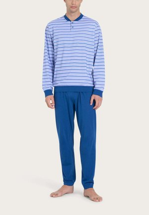 Uomo in piedi a piedi nudi che indossa una maglia a righe blu a maniche lunghe con polsini blu scuro e pantaloni blu scuro tinta unita su sfondo neutro.