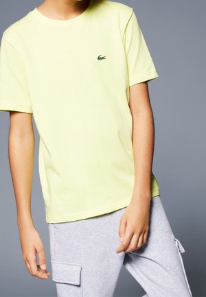 Jeune portant une chemise jaune pâle à manches courtes avec un petit logo crocodile vert et un pantalon cargo gris clair sur un fond uni.