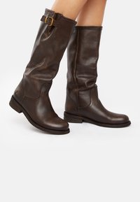Brown leather knee-high boots with a textured finish, featuring a side strap and buckle accent. Low stacked heel and rounded toe design.