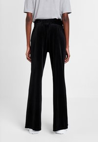 Zwarte fluweelachtige wijde broek met een elastische tailleband, die een verticale ribstructuur heeft en een getailleerde pasvorm biedt. Gestyled met witte schoenen.