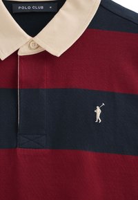 Rot- und marineblau gestreiftes Poloshirt mit beigem Kragen; es hat eine kleine weiße gestickte Figur. Der Stoff ist glatt und gestrickt mit einer Knopfleiste.