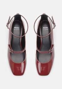 Red patent leather Mary Jane-style heels with a square toe, ankle strap, and buckle detail. Interior features smooth black lining.