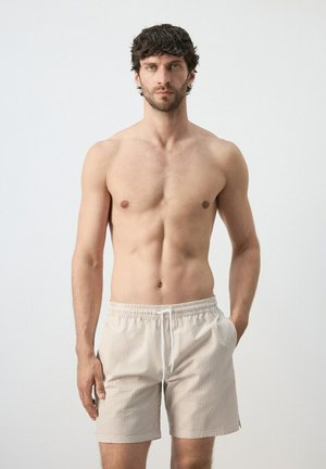 Shirtless man with dark curly hair wearing beige striped drawstring shorts, standing facing forward with one hand in pocket.