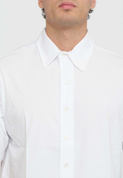 Camicia bianca con colletto classico, chiusura frontale a bottoni e design semplice. Realizzata in tessuto leggero e liscio con una sottile texture.