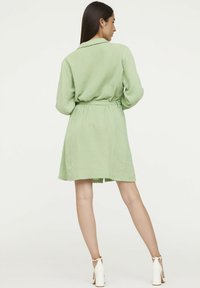 Robe verte à ceinture avec un col, tissu texturé, ourlet à hauteur des genoux et manches longues. Portée avec des chaussures à talons blancs.