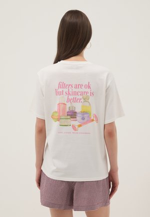 Mujer con cabello largo y castaño que lleva una camiseta blanca con ilustraciones rosas de productos para el cuidado de la piel y texto que promociona el cuidado de la piel sobre los filtros, combinada con pantalones cortos a cuadros.