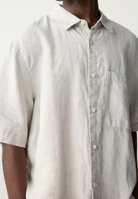 Gros plan d'une personne portant une chemise beige clair à manches courtes avec un bouton-pression et une poche avant, sur un fond uni.