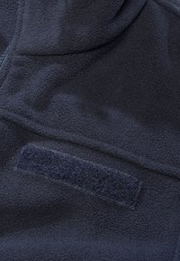 Gros plan d’un tissu polaire bleu marine foncé avec un patch rectangulaire en Velcro et une partie d’une fermeture éclair visible sur le côté gauche.