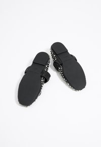 Sandalias planas negras con una textura suave, que presentan una correa ancha adornada con tachuelas metálicas a lo largo de los bordes y un acento de lazo.