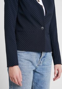 Námořnický modrý blazer s texturou teček, na jedno knoflíkové zapínání, s klopami a bočními kapsami, spárovaný se světle modrými džíny.