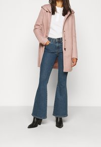 Cappotto rosa con cappuccio sopra una maglietta bianca, abbinato a jeans flare a vita alta blu e stivaletti neri alla caviglia. Tessuto morbido, vestibilità rilassata.