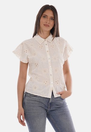 Donna che indossa una camicetta bianca a maniche corte con ricamo floreale a occhiello e jeans blu, in piedi con una mano in tasca.