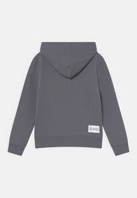 Sudadera gris con capucha, con puños y dobladillo acanalados, que presenta una pequeña etiqueta blanca con "JEANS" en la parte inferior izquierda. Textura suave.