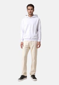 Hoodie en coton blanc avec une poche avant et une capuche à cordon, associé à un pantalon beige et des baskets noires sur un fond neutre.