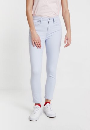 Femme portant un jean slim bleu clair, un haut rose pâle, des chaussettes rouges et des baskets blanches, debout devant un fond blanc.