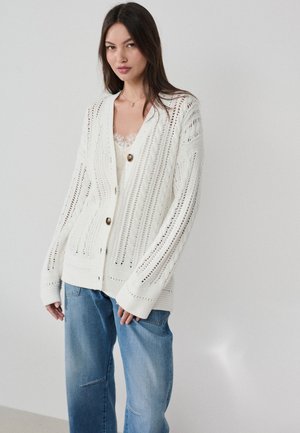 Vrouw draagt een wit opengebreid vest met grote knopen over een kanten top en losse blauwe jeans, staand tegen een effen lichte achtergrond.