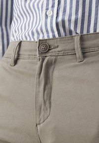 Pantaloni beige con una texture liscia, caratterizzati da una chiusura con bottone e passanti per cintura. Una camicia a righe blu e bianche è parzialmente visibile sopra.