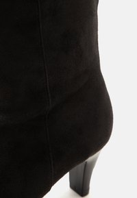 Gros plan de l'arrière d'une chaussure à talon haut en daim noir montrant les coutures et le talon.