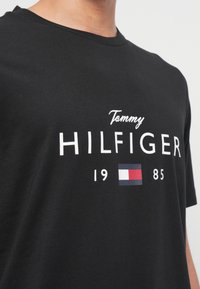 Mustmust kaelustega T-särk, millel on valge tekst "Tommy Hilfiger 1985" ja väike punase, valge ja sinise ristkülikukujuline logo rinnal.
