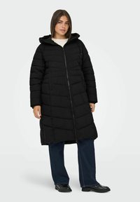 Abrigo acolchado largo negro con capucha, con cierre de cremallera en el frente, diseño acolchado y bolsillos laterales. Lleva puesto sobre unos vaqueros de pierna ancha azules y zapatos negros.