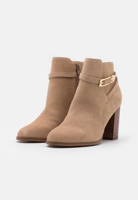 Botines tobilleros de ante beige con cierre de cremallera, una correa decorativa con herrajes dorados y un tacón cuadrado. Textura suave con punta afilada.