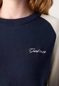 Femme portant un sweat-shirt bleu marine et beige avec "Gant 1949" brodé en blanc sur la poitrine.