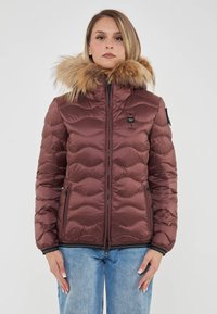 Chaqueta de plumas marrón con capucha forrada de pelo, textura acolchada, cierre de cremallera y bolsillos laterales. Llevada sobre unos vaqueros azules.