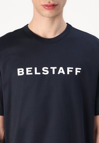Sötétkék pamut póló, környakú szabással és feltűnő fehér "BELSTAFF" felirattal az elején. Sima tapintás, rövid ujjak.