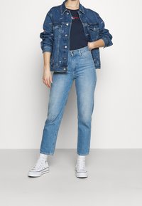 Spijkerjasje met geborduurde accenten, gecombineerd met een donkerblauw shirt, lichtblauwe spijkerbroek en witte hoge sneakers. Casual, getailleerd ontwerp.