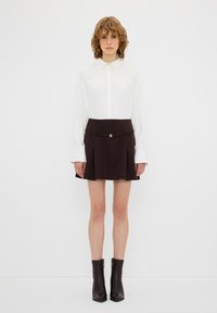 Chemise blanche à boutons avec des manches longues, assortie à une mini-jupe plissée marron avec une ceinture et des accents en doré. Bottines noires.